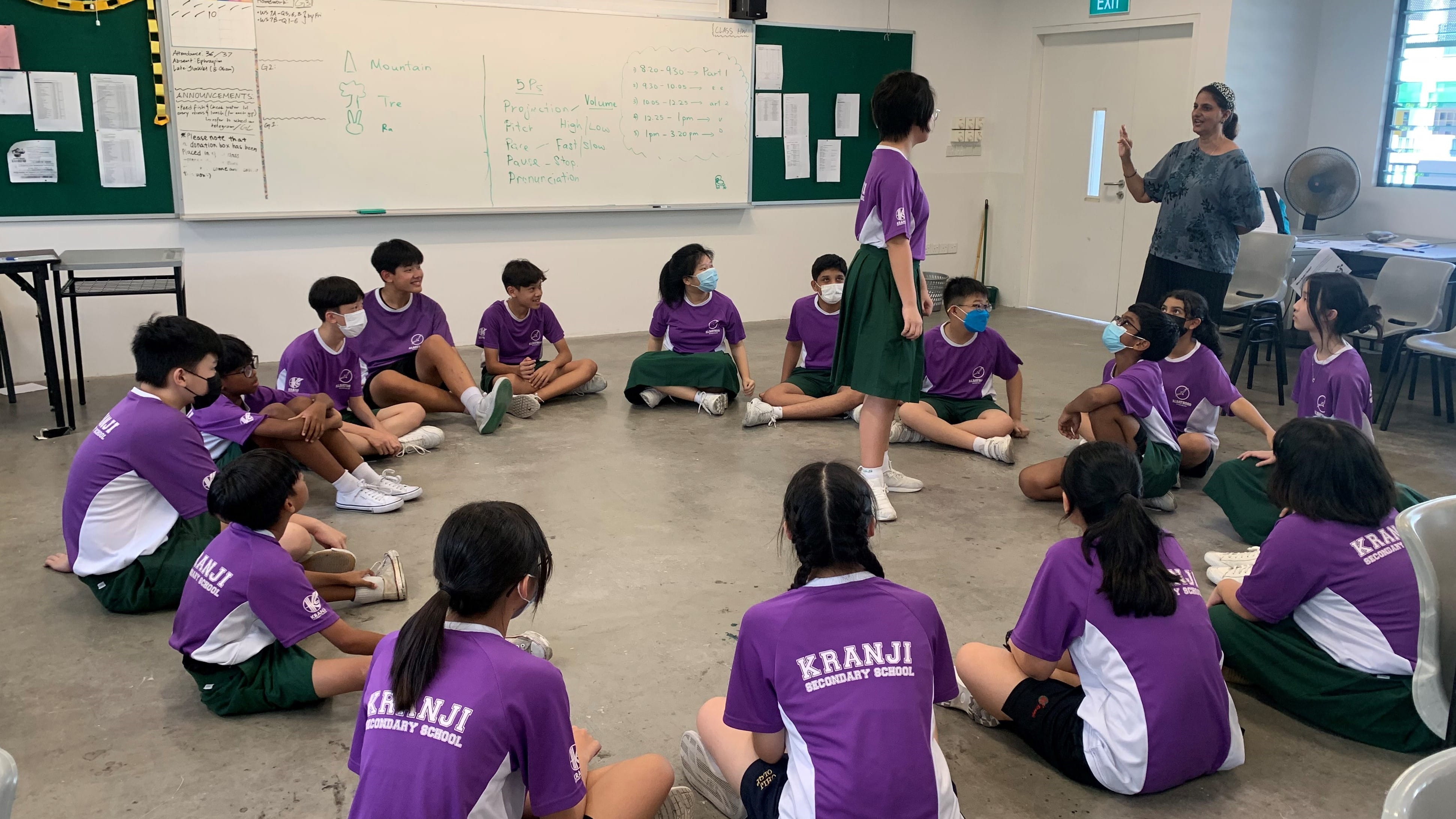 Students in purple Kranji Secondary School uniforms sit in a circle on a classroom floor while a teacher stands nearby.